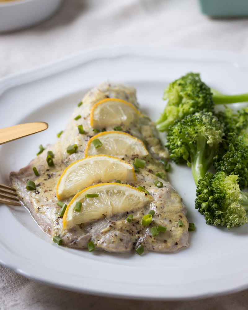 Baked bluefish fillet with sauce served on a white plate with broccoli.
