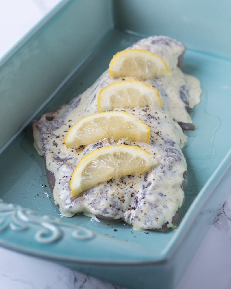 Raw Bluefish fillet in a blue baking dish covered in a creamy sauce. 