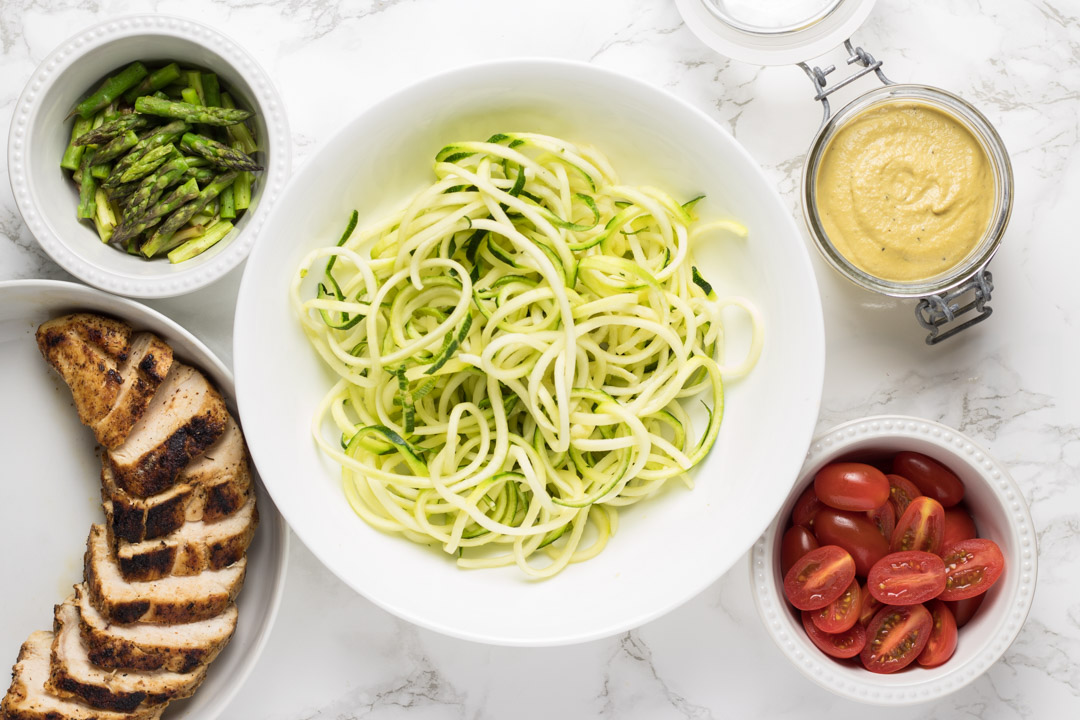 Chicken, asparagus, zoodles, cashew alfredo sauce and cherry tomatoes in white prep bowls