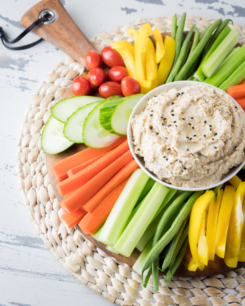 Roasted cauliflower hummus surrounded by colorful vegetables on a textured place mat